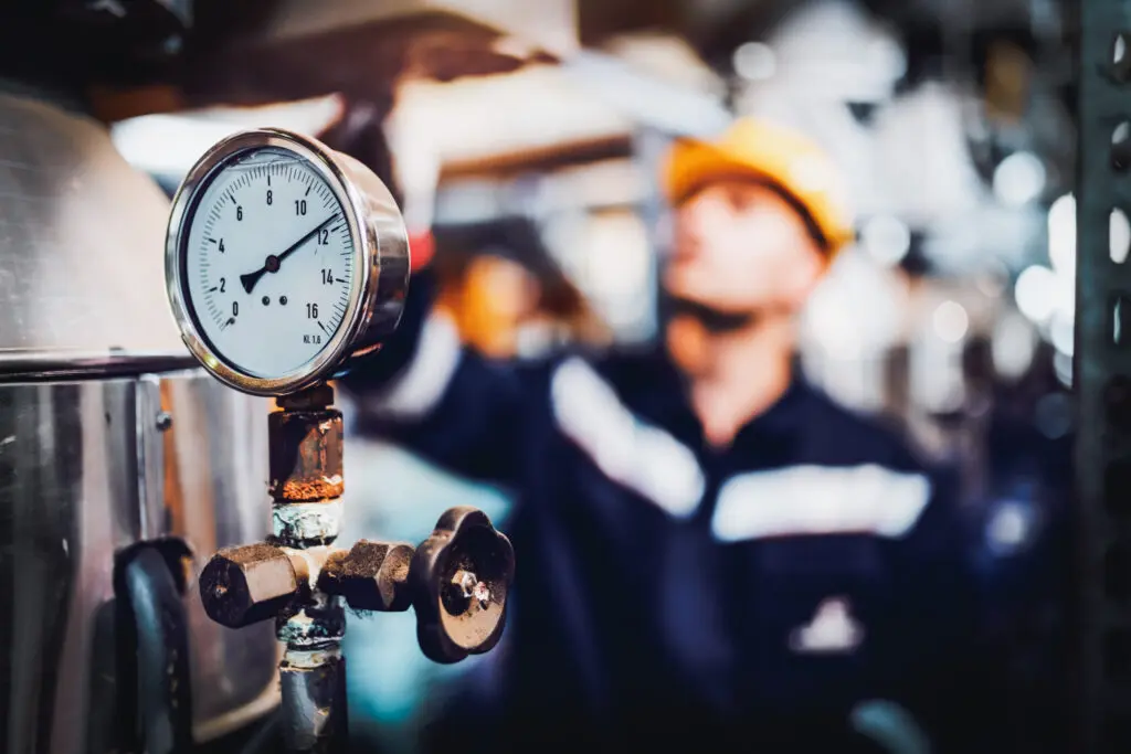 Steam Boiler Maintenance Tips.
Close up of clock on boiler showing pressure on boiler. In background is worker working. Selective focus on clock.